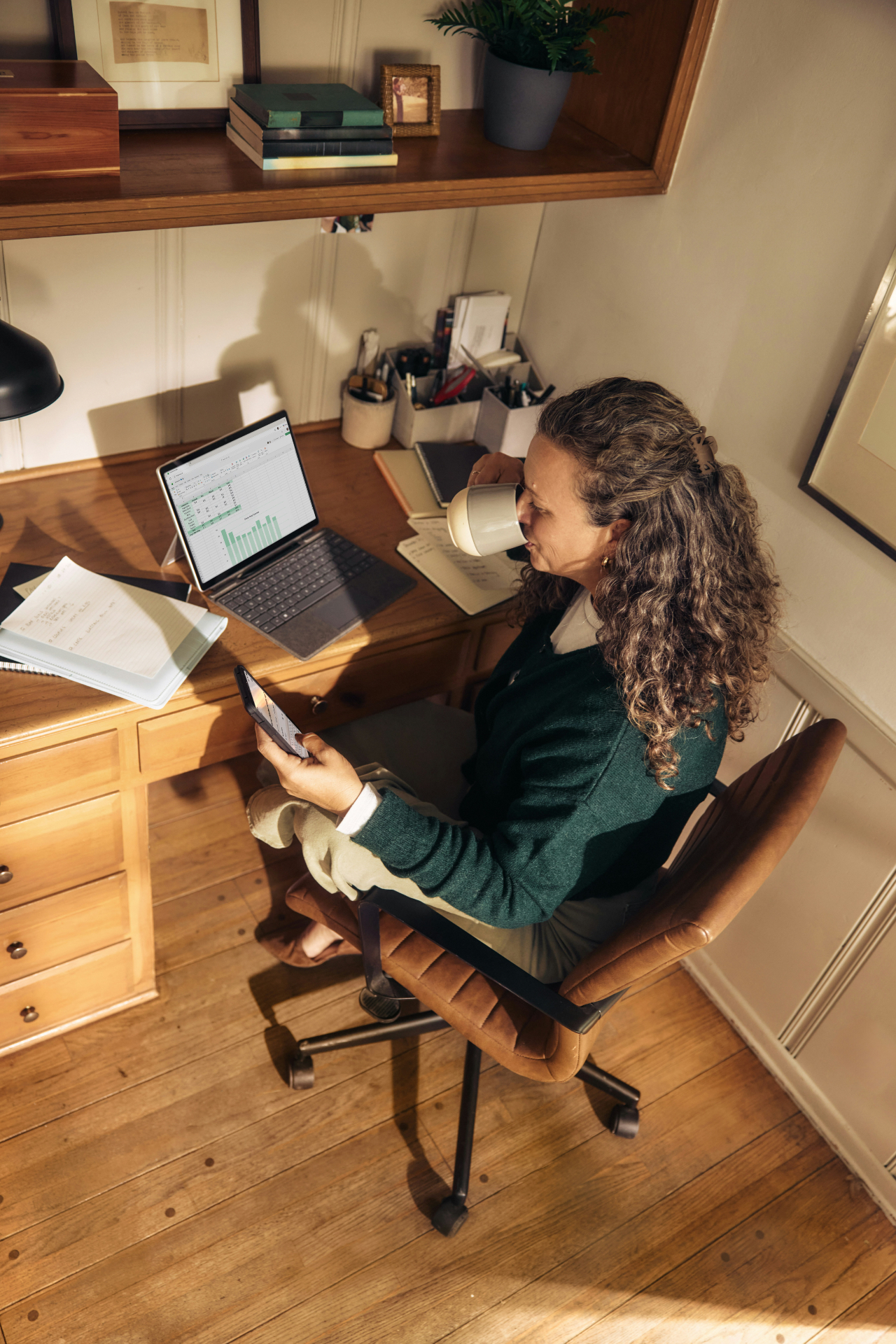 Person am Holzschreibtisch mit Laptop und Tasse Kaffee, prüft konzentriert ihr Smartphone